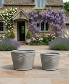 Two gray planters on a stone patio with a garden and house in the background