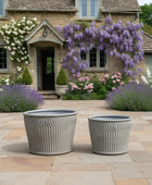 Two gray outdoor planters on a stone patio with a garden and house in the background.