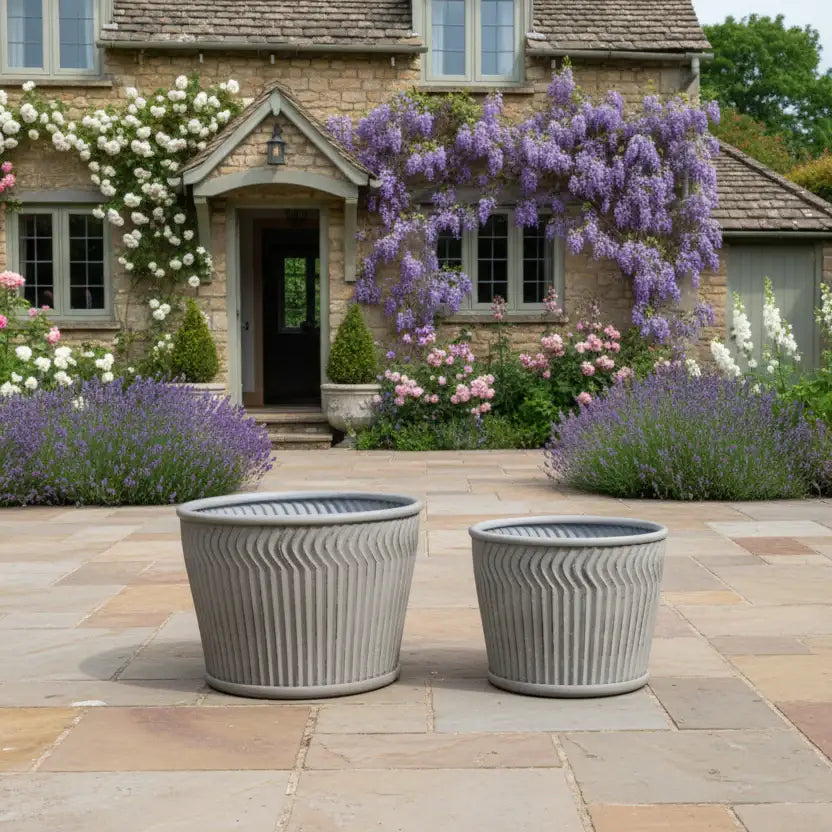 Two gray outdoor planters on a stone patio with a garden and house in the background.