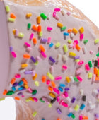 A close-up of a donut or pastry with pale pink icing and scattered rainbow sprinkles, photographed against a white background.