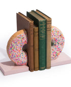 A pair of novelty donut-shaped bookends holding up a row of vintage-style books on a clean white background. The bookends are light pink with raised donut shapes covered in multicolored sprinkles, and the books in between add brown, green, and tan tones.