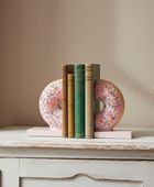 A pair of pink donut-shaped bookends with colorful sprinkles holding a set of four books on a distressed white wooden console table, with a potted green plant to the right against a beige wall.