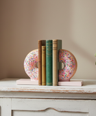Two pink donut-shaped bookends with colorful sprinkles and light pink bases holding a row of books on a distressed white wooden table against a beige wall.