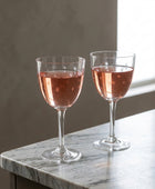 Two clear wine glasses on a white tablecloth, one filled with pink wine and one empty with etched star details, with peaches in the background.