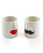 Two white ceramic novelty cups shaped like faces, one with red lips and one with a black mustache, on a white background.