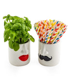 Two white ceramic cups shaped like stylized faces, one with red lips holding a green basil plant and the other with a black mustache holding a set of colorful striped paper straws, on a white background.