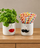 Two white ceramic cups shaped like faces sit on a wooden countertop. The left cup has a red lips design and holds a green basil plant, while the right cup has a black mustache design and holds a set of colorful striped paper straws.