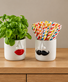 Two white ceramic cup holders shaped like faces sit on a wooden surface against a beige wall. The left holder has a red lips design and contains a green basil plant.