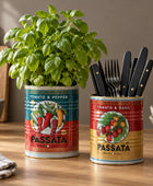 Two vintage-style metal canisters with colorful tomato-themed labels sit on a countertop. One canister holds a leafy green herb plant, and the other holds two gold-toned metal spoons.