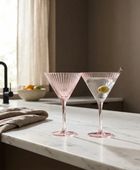 Two pink fluted glass martini glasses on a white marble kitchen counter, with one empty and one filled with a clear drink and an olive garnish.