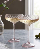 Two clear champagne coupe glasses with a twisted, textured pattern and a subtle pink tint, shown on a light olive green tablecloth with a white curtain backdrop.