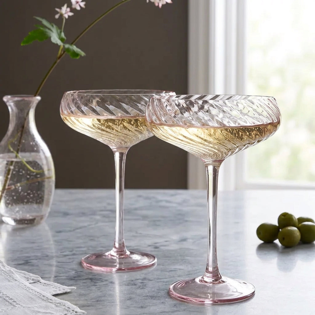 Two clear champagne coupe glasses with a twisted, textured pattern and a subtle pink tint, shown on a light olive green tablecloth with a white curtain backdrop.