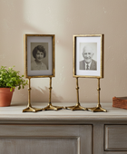 Two gold-framed photographs on stands with a plant and book on a wooden surface.