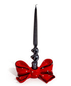 A black taper candle with a spiral twist base, set in a glossy red bow-shaped holder with white polka dots, on a plain white background.