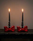 Two black spiral taper candles with bright flames, set in matching black holders decorated with glossy red bows, placed on a dark rustic table against a plain dark wall.