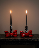 Two black taper candles in spiral holders with red bow-shaped bases, displayed on a dark wooden table against a warm brown wall.