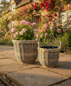 Two wicker planters with flowers on a stone patio in front of a house with floral decorations.
