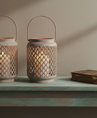 Two rustic metal lantern candles with wire handles sit on a distressed teal table next to an old book against a warm beige wall with soft shadows.