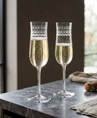 Two clear champagne flutes filled with pale golden liquid, set on a white fabric surface with a small sprig of greenery and a cork in the background.