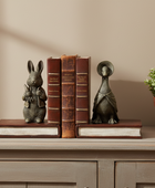 Two decorative bookends shaped like a rabbit and a duck sitting on a shelf next to three vintage-style brown books and a potted green plant against a beige wall.