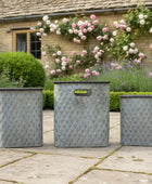 Three metal planters on a patio with a garden and house in the background