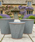 Three gray metal planters on a stone patio with a house and flowers in the background