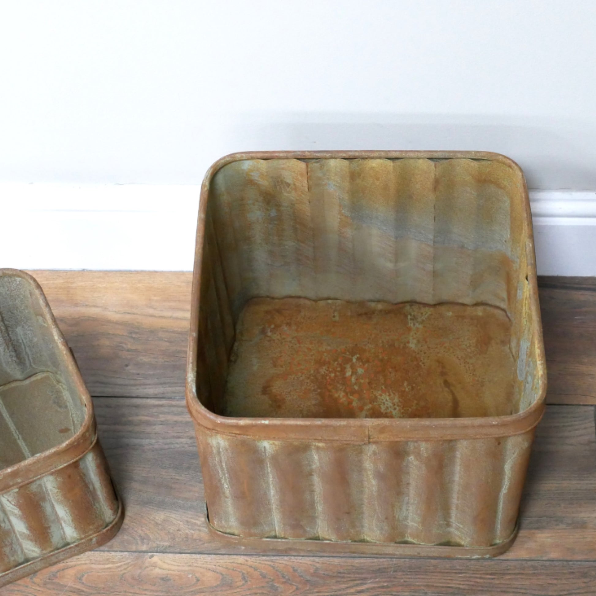 Two rustic metal bins on a wooden floor with a white wall background.