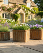 Three wooden planters with flowers on a stone patio in front of a house with garden.