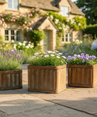 Three wooden planters with flowers on a patio in front of a house with garden.