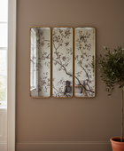 Three vertical rectangular mirrors with gold frames hang on a taupe wall in a styled room, featuring a delicate branch and bird pattern. A potted olive tree is visible on the right, and a window frame is on the left.
