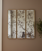 Three vertical rectangular mirrors with gold frames hang side by side on a warm taupe wall. The mirrors feature a vintage-style floral branch pattern in muted brown and gray tones on a distressed off-white background.