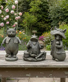Three stone statues of a cat, frog, and mouse on a wooden bench with a garden background.