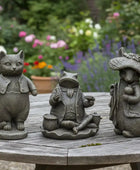 Three garden statues of a cat, frog, and mouse on a wooden table with a garden background.