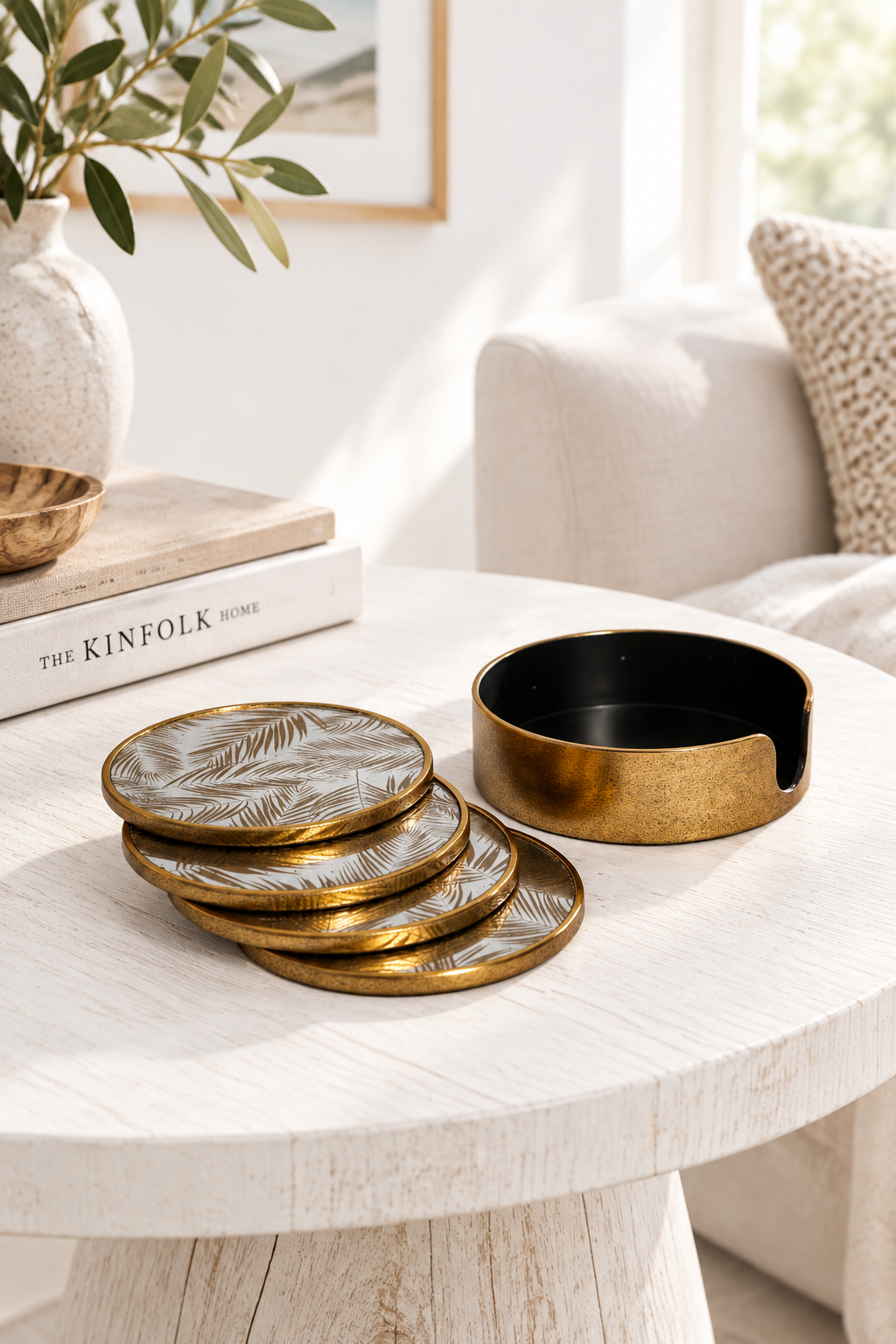 A set of four round coasters with a gold-toned metallic border and a white base featuring a gold palm leaf pattern, displayed on a light-colored wooden table next to a matching gold and black cup in a bright, modern living room setting.