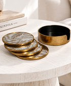 A set of round coasters and a matching cup holder with a brushed gold finish and a leaf pattern, styled on a white wooden table next to books.