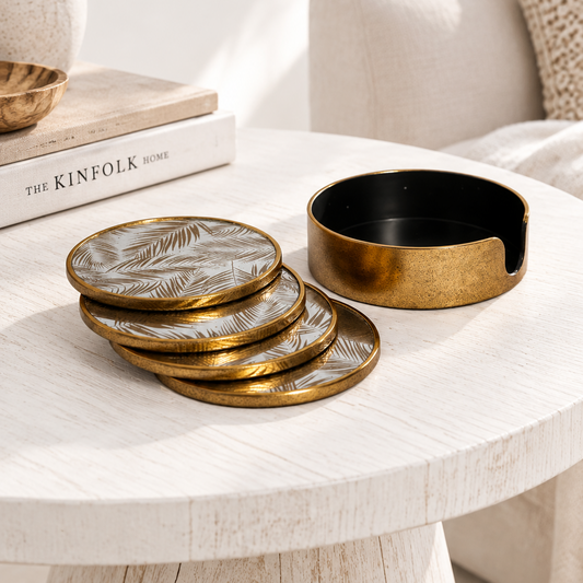 A set of round coasters and a matching cup holder with a brushed gold finish and a leaf pattern, styled on a white wooden table next to books.