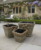 Three wicker planters with black liners on a stone patio in front of a stone building with flowers.