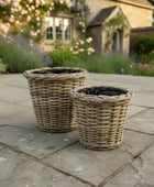 Two wicker planters on a stone patio with a garden and house in the background.