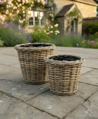 Two wicker planters on a stone patio with a garden and building in the background