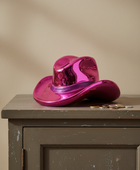 A shiny metallic magenta pink cowboy hat resting on a distressed gray wooden cabinet against a beige wall.