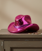 A shiny metallic magenta pink cowboy hat container with a coin slot on the brim, resting on a distressed gray wooden cabinet against a beige wall.