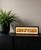 A rectangular illuminated sign reading "SHOWTIME" in raised gold letters on a warm cream background, placed on a dark cabinet beside a potted fern in a real indoor setting.
