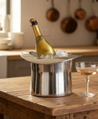 Bottle of champagne in a metal ice bucket on a wooden table with a glass in a kitchen setting.