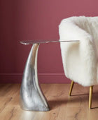 Silver side table with a clear glass top next to a white fur-covered chair against a red wall.
