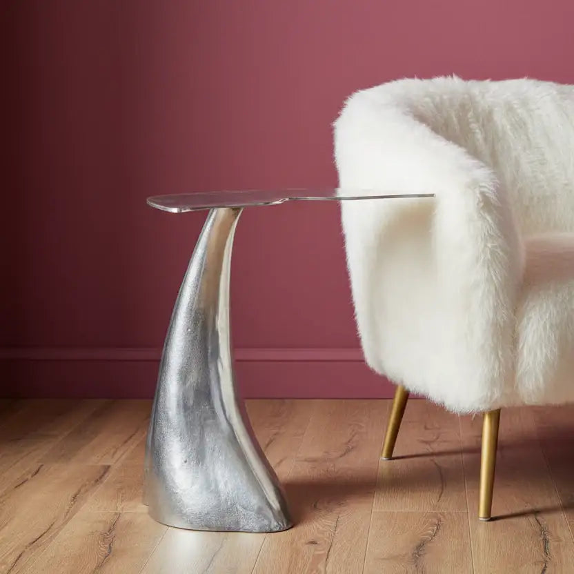 Silver side table with a clear glass top next to a white fur-covered chair against a red wall.