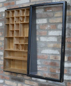 A wall-mounted wooden display cabinet with multiple small open compartments on the left and a large black-framed glass door panel on the right, shown against a brick wall.