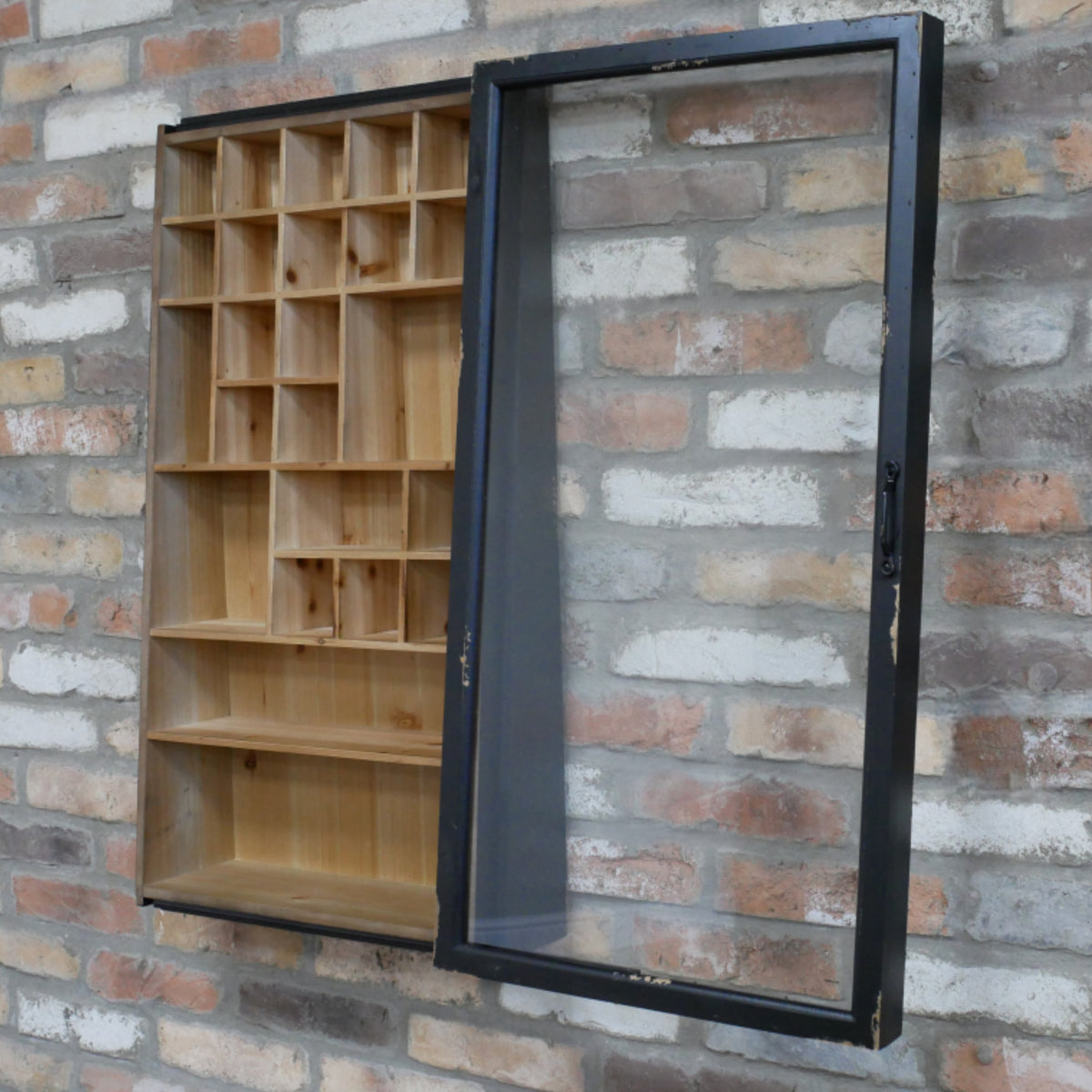 A wall-mounted wooden display cabinet with multiple small open compartments on the left and a large black-framed glass door panel on the right, shown against a brick wall.