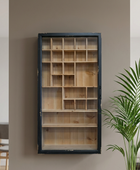 A tall, narrow wall-mounted wooden display cabinet with a dark navy frame and multiple small interior compartments, shown against a warm taupe wall with a green plant at the right edge.