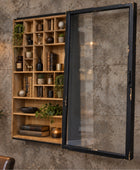 A wall-mounted wooden bookshelf with an attached black-framed glass door, styled with books, vases, and plants against a textured concrete wall.
