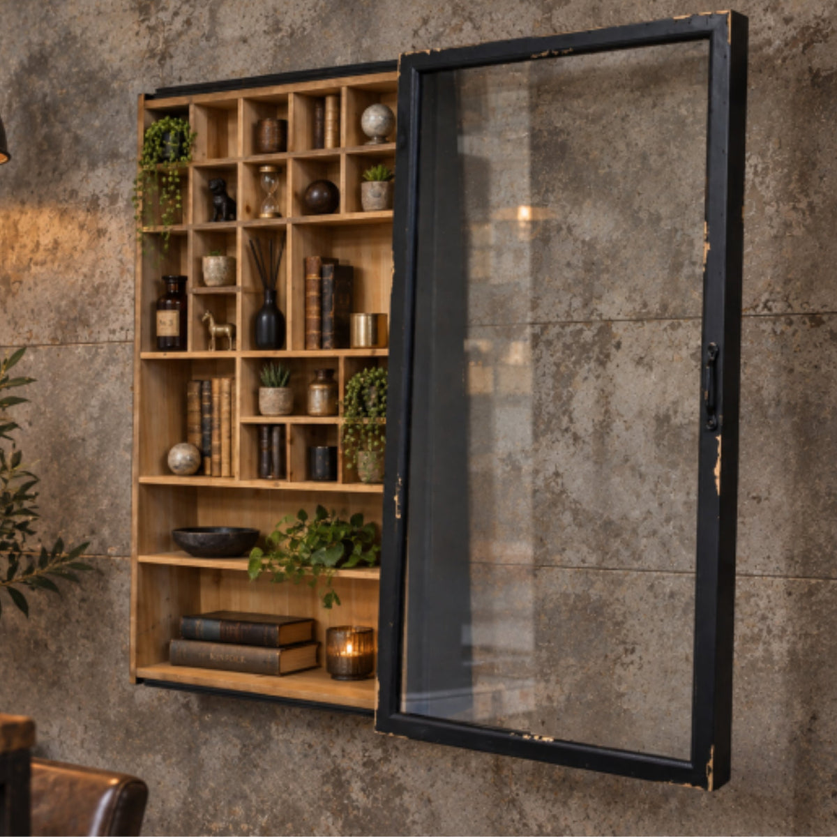 A wall-mounted wooden bookshelf with an attached black-framed glass door, styled with books, vases, and plants against a textured concrete wall.